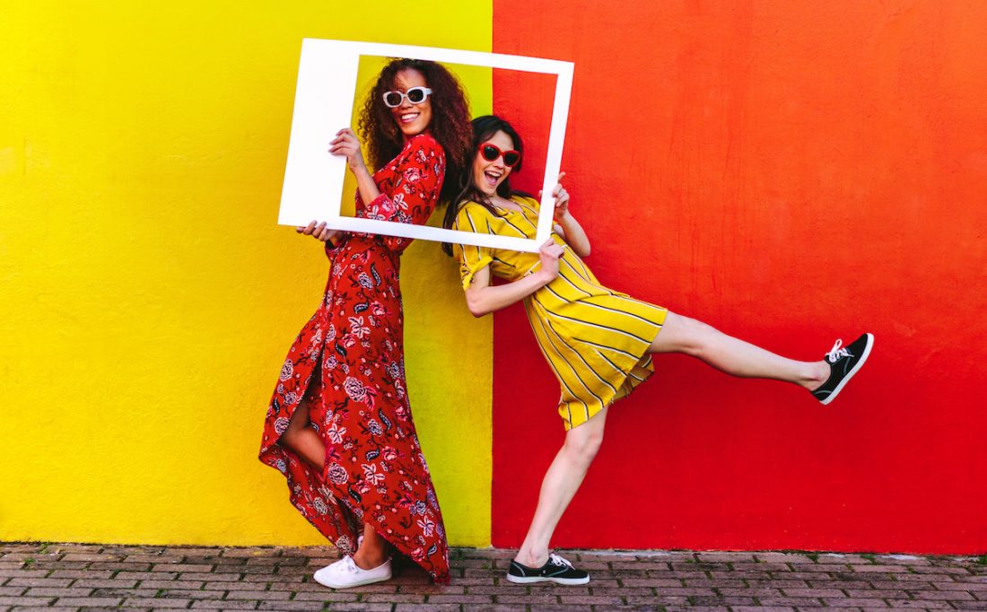 Two women friends with blank photo frame standing against colored wall outdoors. Female travelers posing at camera with empty picture frame.