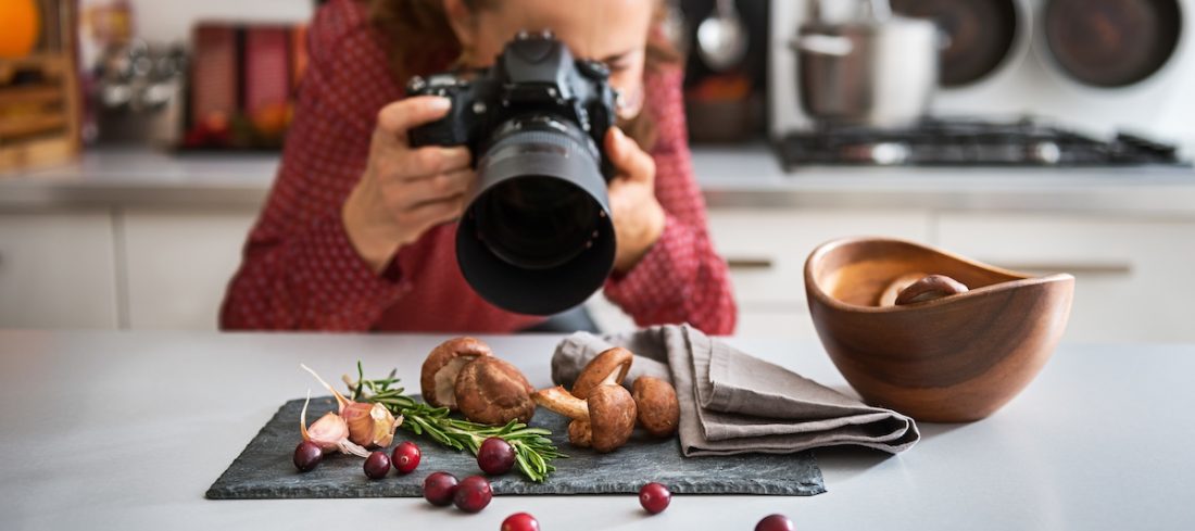 A woman food photographer in the background leans down to take a close-up, in a modern kitchen, of autumn fruits and vegetables - mushrooms, garlic, rosemary, and cranberries.